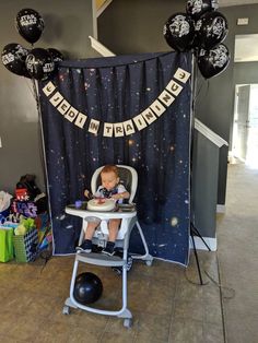 a baby sitting in a high chair next to some black and white balloons on the wall