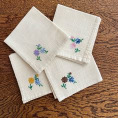 four white napkins with embroidered flowers on them sitting on a wooden table top next to each other