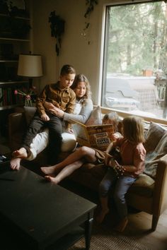 three people sitting on a couch in front of a window with a book open to read