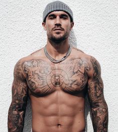 a shirtless man with tattoos on his chest and hands behind his back, standing in front of a white wall