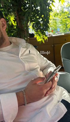 a man sitting in a chair looking at his cell phone while wearing a white shirt