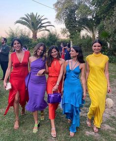 four women in different colored dresses are walking on the grass with palm trees behind them