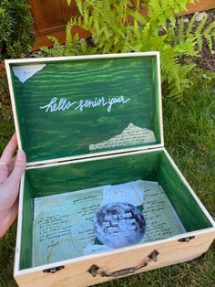 someone is holding an open wooden box with writing on it and there is a plant in the background