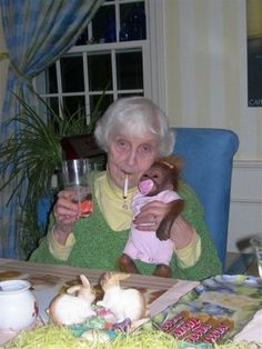 an elderly woman holding a teddy bear and drinking wine at a table with other stuffed animals