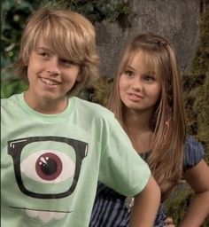 two young children standing next to each other in front of a wall with moss growing on it