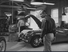 a man standing next to a car in a garage with other men working on it