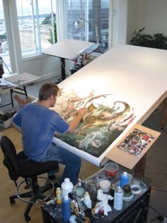 a man sitting in front of a large painting on top of a white table next to a black chair