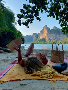 a woman laying on top of a beach next to a basket and book in her hand