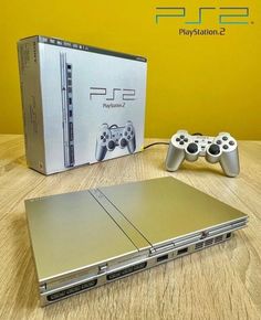 a silver sony playstation 2 console and two game controllers on a wooden table with a yellow wall in the background