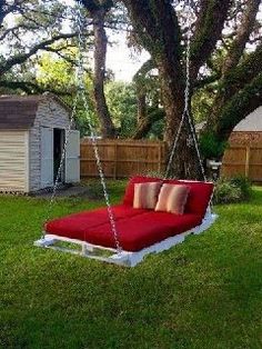 a red swing bed hanging from a tree in a backyard with green grass and trees