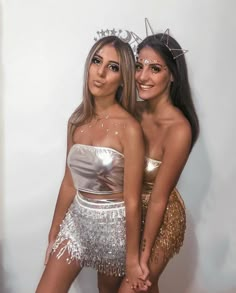 two beautiful young women standing next to each other in front of a white wall wearing silver and gold outfits