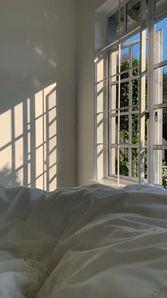 an unmade bed with white sheets and pillows in front of two windows that look out onto the trees