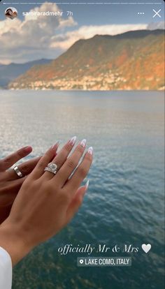 two hands holding each other over water with mountains in the background and clouds in the sky