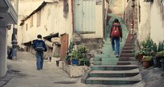 two people are walking up the stairs in an alleyway with potted plants on either side