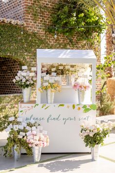 a small white stand with flowers and plants on it in front of a brick building