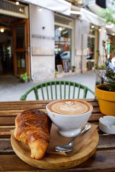 a cup of coffee and croissant sit on a wooden table in front of a cafe