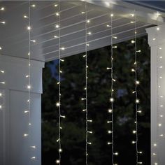 some lights are hanging from the side of a porch with white curtains and windows in the background