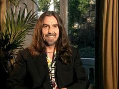 a man with long hair sitting in front of a window and smiling at the camera