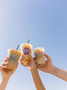 three people hold up cups with drinks in them and the sky is blue behind them