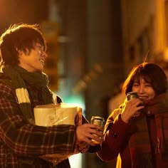 two people standing next to each other holding drinks and looking at their cell phones in the street