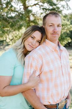 a man and woman standing next to each other in front of some trees with their arms around each other