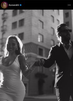 a man and woman holding hands while walking down the street in front of a tall building