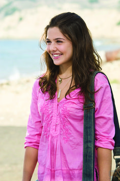 a woman in pink shirt and jeans holding a black bag smiling at the camera while standing on beach