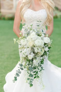 a woman in a wedding dress holding a bridal bouquet with white flowers and greenery