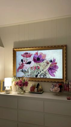 a painting hanging on the wall above a dresser with flowers in vases and other items