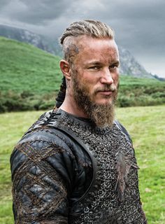 a man with long hair and beard standing in front of a green field on a cloudy day