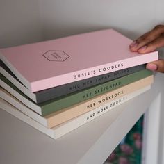 a person holding a stack of books on top of a white shelf in front of a wall