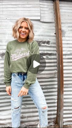 a woman standing in front of a metal wall wearing a green sweatshirt and ripped jeans