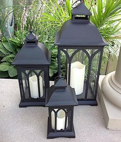 three black lanterns sitting on top of a cement floor next to plants and potted plants