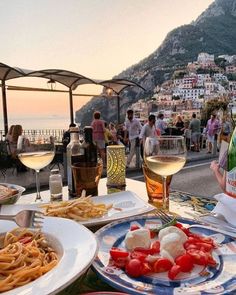 plates of food and glasses of wine on a table overlooking the ocean with people standing around