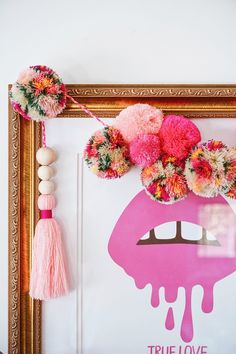 a pink poster with pom - poms hanging from it's sides and a painting on the wall behind it