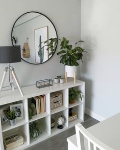 a white shelf with books and plants on it in front of a round mirror above