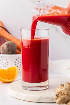 a person pouring juice into a glass on top of a table with carrots and an orange