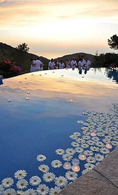 people are sitting on the edge of a swimming pool with flowers in the water and candles floating around