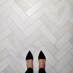 a woman's feet in black shoes standing on a white tile floor with herringbone pattern