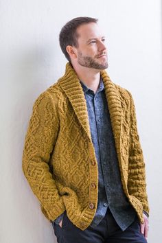 a man standing against a wall with his hands in his pockets, wearing a yellow cardigan