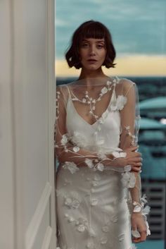 a woman in a white dress standing next to a wall and looking at the camera