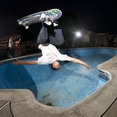 a skateboarder does a trick in an empty pool at night while another man watches