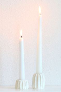 two white candles sitting next to each other on top of a table in front of a wall