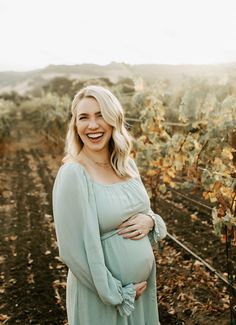 a pregnant woman smiles while standing in a vineyard