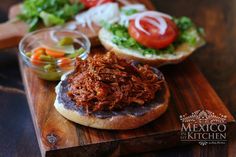 a pulled pork sandwich with lettuce, tomato and pickles on a wooden cutting board