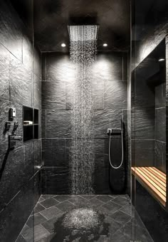 a shower head in the middle of a black tiled bathroom with wooden bench under it