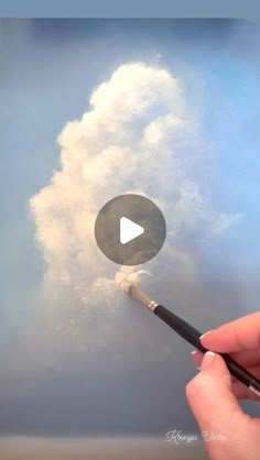 a hand holding a paintbrush with clouds in the sky behind it and a video play button