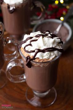 two cups filled with hot chocolate and whipped cream on top of a wooden table next to a christmas tree