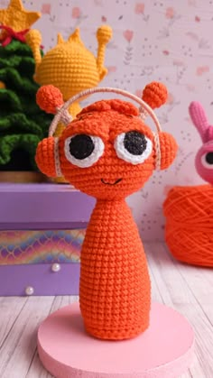 an orange crocheted toy with big eyes and headphones on top of a pink base