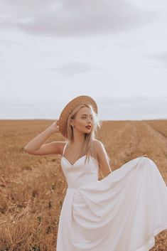 a woman in a white dress and straw hat standing in a field with her arms behind her head